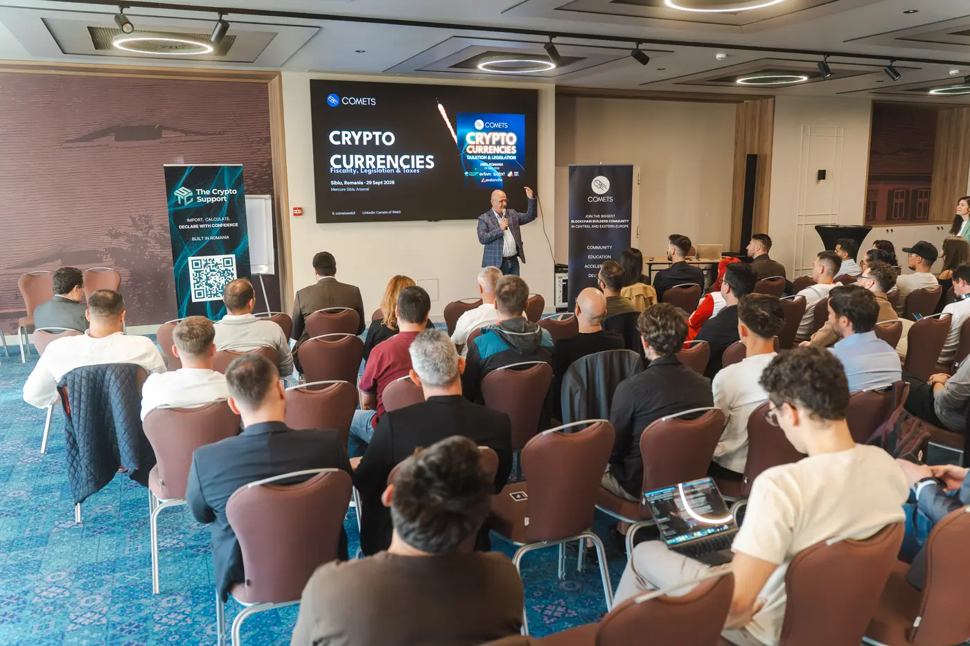 Sala plină la COMETS Crypto Currencies, cu un vorbitor pe scenă în fața ecranului cu numele evenimentului.